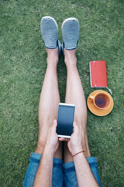 Adam akıllı telefon kullanarak ve bir çim üzerinde kahve / çay içme.