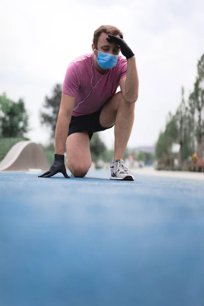 Sportsman with medical mask and gloves, smartphone and earbuds working out, jogging in urban surroundings. Exhausted man from jogging and medical mask usage. Trouble with breathing.