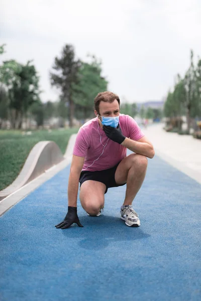 Sportsman with medical mask and gloves, smartphone and earbuds working out, jogging in urban surroundings. Exhausted man from jogging and medical mask usage. Trouble with breathing.