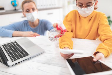 Tablet ve laptop sterilize ediliyor. Virüsler, grip ve salgın zamanında evde eğitim, evde eğitim, özel öğretmen / koruyucu maskeli anne ile eğitim gören bir çocuk.