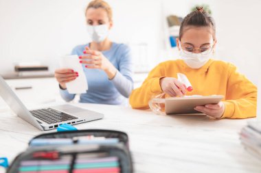 Tablet ve laptop sterilize ediliyor. Virüsler, grip ve salgın zamanında evde eğitim, evde eğitim, özel öğretmen / koruyucu maskeli anne ile eğitim gören bir çocuk.