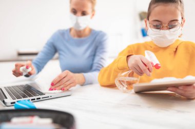 Tablet ve laptop sterilize ediliyor. Virüsler, grip ve salgın zamanında evde eğitim, evde eğitim, özel öğretmen / koruyucu maskeli anne ile eğitim gören bir çocuk.