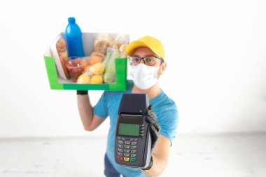 Koruyucu maskeli teslimatçı. Bakkaliye ve POS 'la dolu bir çanta tutuyor..