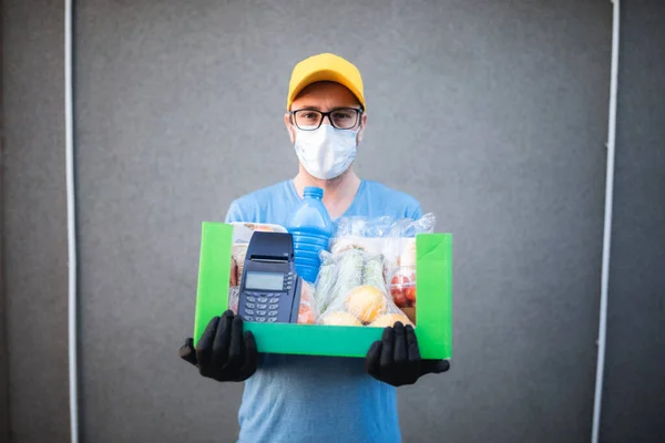 Delivery guy with protective mask holding box / bag with groceries and POS for contactless payment.