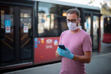 Tıbbi koruyucu maskeli ve eldivenli bir adam otobüs durağında toplu taşıma bekliyor ve akıllı telefon kullanıyor..