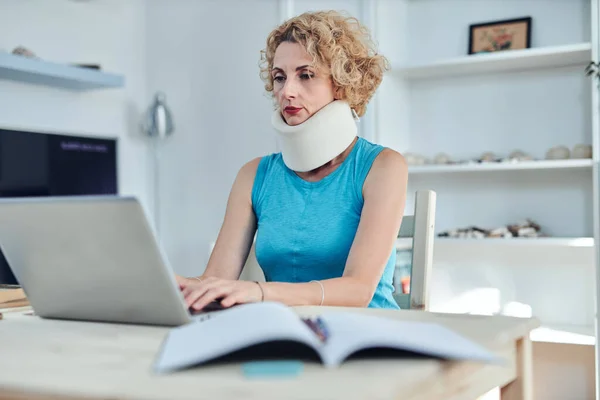 Woman with neck / cervical collar and neck / spinal injury working at home on a laptop.