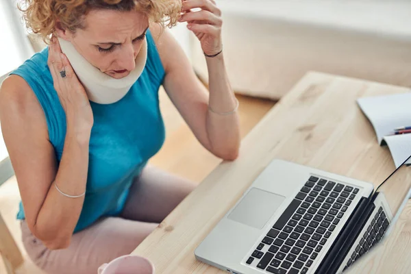 Woman with neck / cervical collar and neck / spinal injury working at home on a laptop.