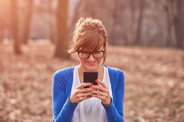 Sonbahar parkında akıllı telefon kullanan genç bir kadın..