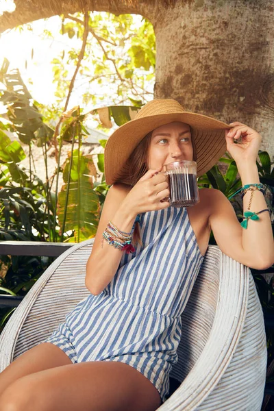 Young caucasian woman sitting on a patio and drinking coffee / tea in summertime season.