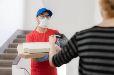 Koruyucu maskeli teslimatçı elinde pizza kutusu ve kapıda kontak kartı olan bir adam..