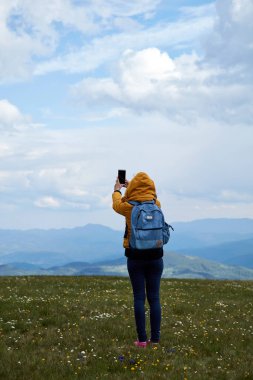Bir kadın cep telefonunu geniş bir çayırda ve dağ manzarasında kullanıyor..