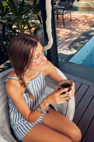 Young caucasian woman sitting on a patio and drinking coffee / tea in summertime season.