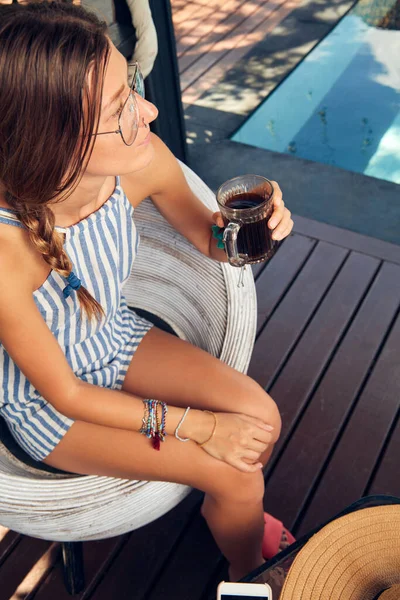 Young caucasian woman sitting on a patio and drinking coffee / tea in summertime season.