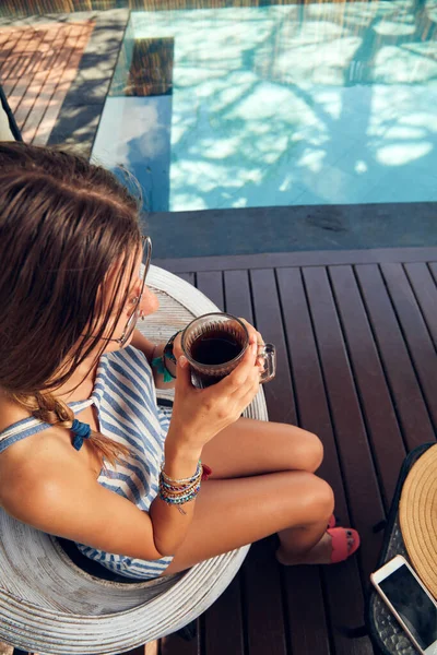 Young caucasian woman sitting on a patio and drinking coffee / tea in summertime season.