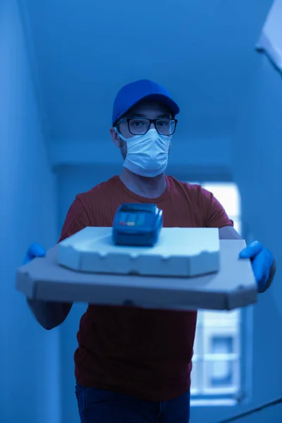 Delivery guy with protective mask holding pizza box and contactless POS terminal at the door.