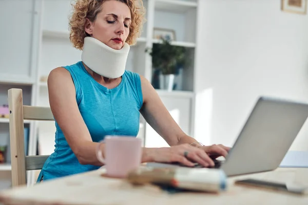 Woman with neck / cervical collar and neck / spinal injury working at home on a laptop.