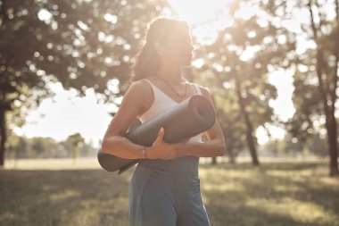 Gün batımında parkta yoga minderi tutan kadın..