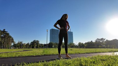 Genç bir kadın güneşli yaz gününde şehir parkında egzersiz ve koşu için ısınıyor..