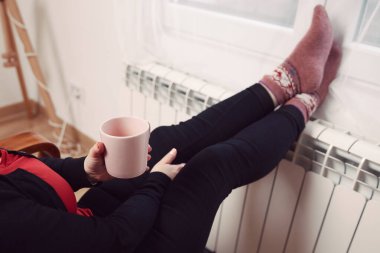 Soğuk bir kış gününde, sıcak çay içen bir kadın, çoraplarıyla evde ısıtma radyatöründe çoraplarını giyiyor..