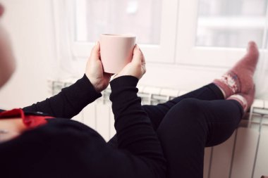 Soğuk bir kış gününde, sıcak çay içen bir kadın, çoraplarıyla evde ısıtma radyatöründe çoraplarını giyiyor..