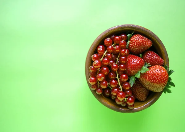 Bowl of red mixed berries on green background. Berry mix. Delicious fresh ripe red currant and strawberry in a brown bowl, plate, dish. Isolated berry mix with copy space. Space for text. Top view.
