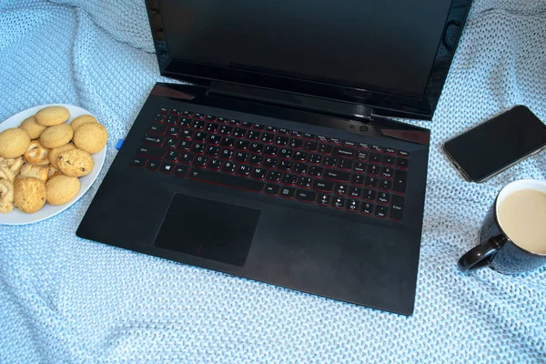 Black laptop, mobile, cookies and cup of coffee on turquoise blanket. Freelance work at home. Winter mood.