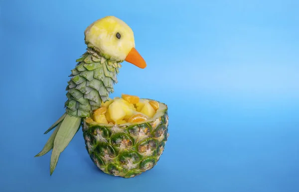 Sliced fruits in pineapple bowl decorated with parrot carved from a pineapple on blue light background with copy space. Food art, food humor and carving. Pineapple\'s parrot bird