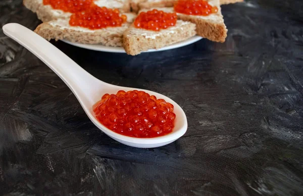 White spoon with red caviar and plate of triangle sandwiches on dark gray or black background. Red caviar sandwiches. Healthy food and snack.