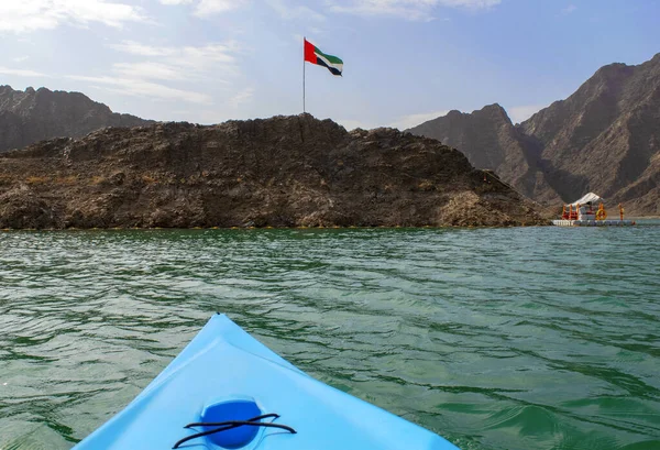 United Arab Emirates flag waving on top of a hill in the Middle of Hatta lake with blue in color kayak on a foreground. Beautiful UAE flag. Green dam for kayaking.