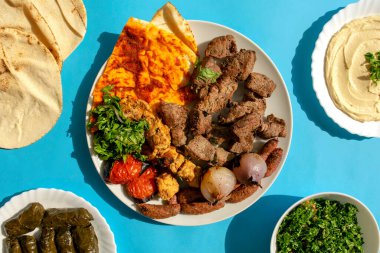 Ünlü geleneksel Arapça, Türkçe, İsrail yemekleri. Izgara tavuk, kuzu, sığır eti, sosis, sebze, pita ekmeği, tahini sosu, mahshi, doldurulmuş asma yaprağı, mavi arka planda tabbouleh salatası, üst manzara.