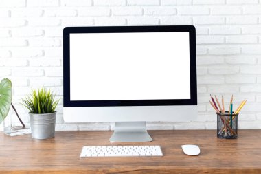 Boş beyaz ekranlı bilgisayar ve ahşap bir masada ofis malzemeleri olan çalışma alanı.