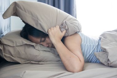 Yatakta yatan genç bir kadın yastıkla başını ve kulaklarını kaplayan sesten muzdarip ve tatsız bir yüz ifadesi takınıyor. Erken kalktım. Yeterince uyuyamadım.