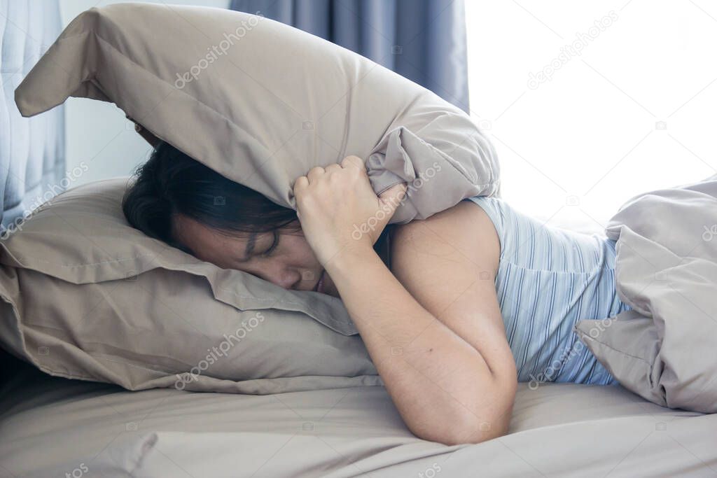 Young woman lying in bed suffering from sound covering head and ears with pillow making unpleasant face. early wake up not getting enough sleep