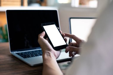 İşadamı elinde boş beyaz ekranlı akıllı telefon ve kafeteryadaki ahşap masada bilgisayar tutuyor.