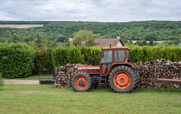 Red tractor parked in a garden in front a stack of cut wood. Countryside landscape.