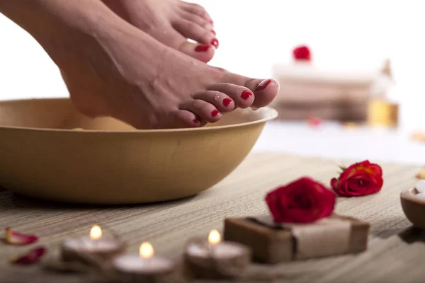 Female feet with drops of water, spa bowl, towels, flowers, candles, sea salt and bottle with essential oil on white background. Foot spa concept.