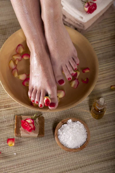 Female feet with spa bowl, flowers, sea salt and bar soap on wooden background. Foot spa concept.