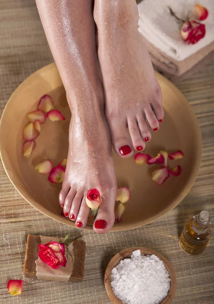 Female feet with spa bowl, flowers, sea salt and bar soap on wooden background. Foot spa concept.