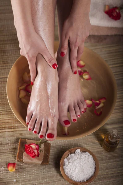 Female feet and hands with spa bowl, flowers, sea salt and bar soap on wooden background. Foot spa concept.