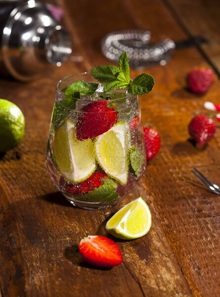 Summer cold drink with strawberries, mint, lime and ice in glass on wooden bar counter. Closeup of cocktail with fresh berry fruits.