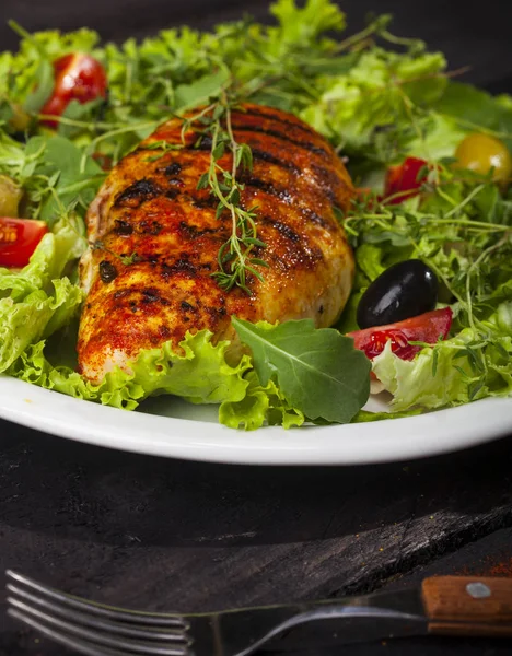 Grilled chicken fillet with fresh lettuce, thyme, cherry tomatoes, and olives in a plate on a black background