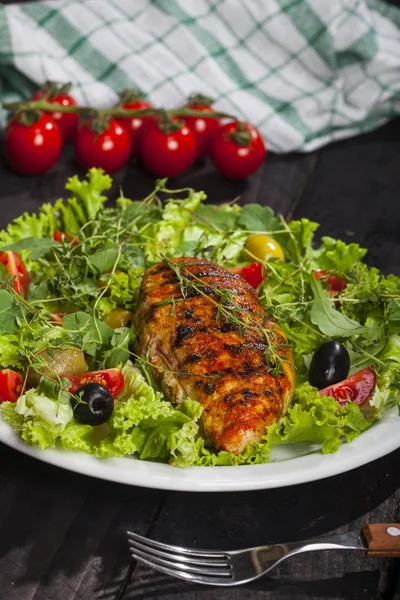 Grilled chicken fillet with fresh lettuce, thyme, cherry tomatoes, and olives in a plate on a black background