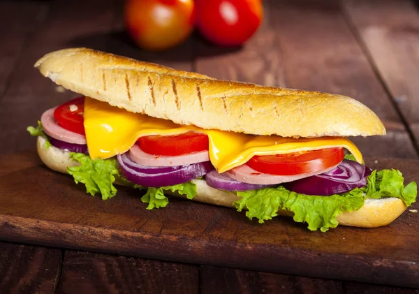 Closeup of a baguette sandwich with ham, cheese, lettuce, tomatoes and onion on a cutting board. Long subway sandwich on a dark background.