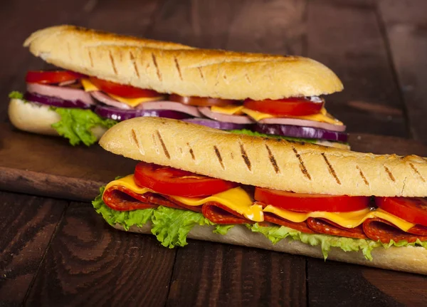 Two baguette sandwiches with salami, ham, cheese, lettuce, tomatoes and onion on a cutting board. Long subway sandwiches on a dark background.