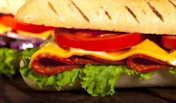 Two baguette sandwiches with salami, ham, cheese, lettuce, tomatoes and onion on a cutting board. Long subway sandwiches on a dark background.