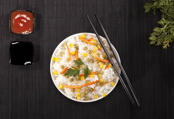 Cooked white rice with vegetables in a bowl with sauces and chopsticks on the wood black bamboo background.