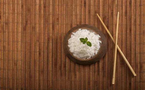 Vařená bílá rýže v dřevěné misce s hůlkami na dřevo hnědé bambusové pozadí. — Stock fotografie