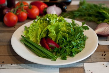 Tahta bir masada beyaz bir tabakta çeşitli taze dereotu, maydanoz, soğan ve biber. Vejetaryen yemeği.