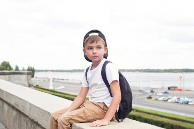 Beyaz tişörtlü ve sırt çantalı Avrupalı bir çocuk parkta oturuyor ve ileriye bakıyor.