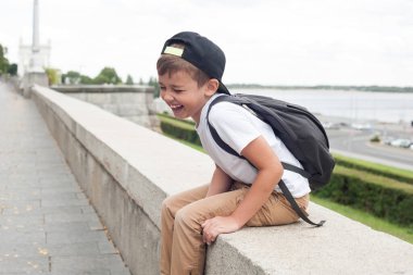 Beyaz tişörtlü ve sırt çantalı Avrupalı bir çocuk oturmuş mutluluktan gülüyor.